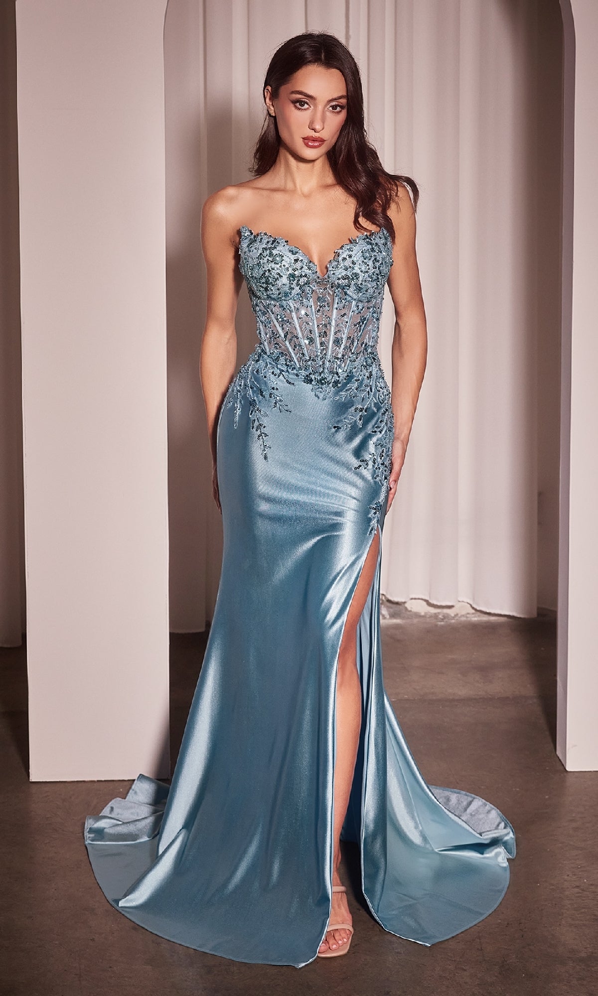 A woman stands indoors in the Strapless Long Prom Dress Ladivine KV1112, light blue with a form-fitting satin skirt, sheer bodice, floral embellishments, and a high slit.